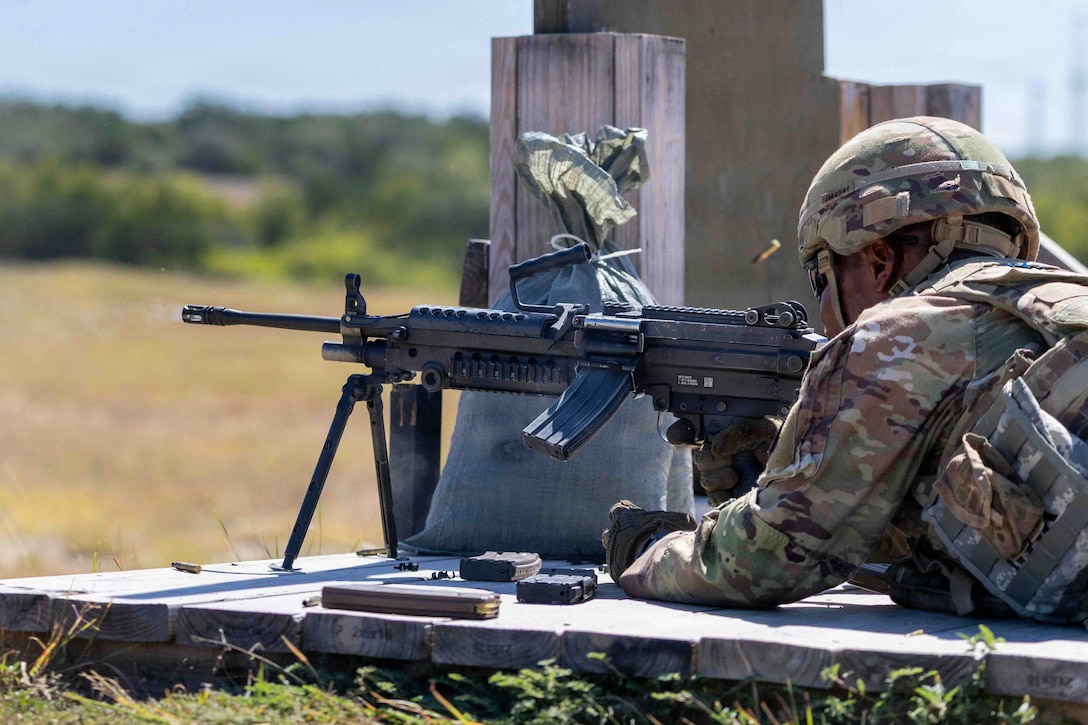 A soldier in a field lies on wooded boards behind a machine gun and fires it with one hand. A shell casing is in the air and a barricade is to the right.