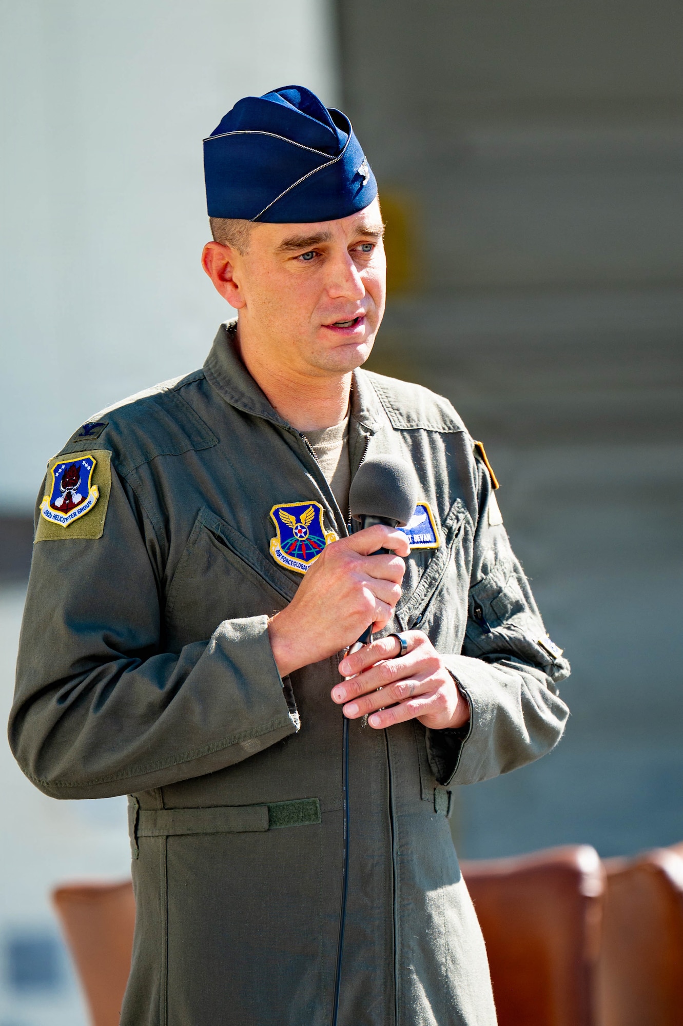 Col. Bryant Bevan Speaking while holding a microphone in front of a helicopter hangar building.