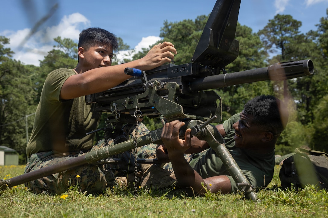 Two Marines in green T-shirts and camouflage pants assemble a grenade launcher on a field on a clear, sunny day.