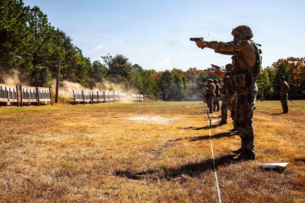 Service members standing in a row on a field fire pistols at targets as a couple of others observe behind them.