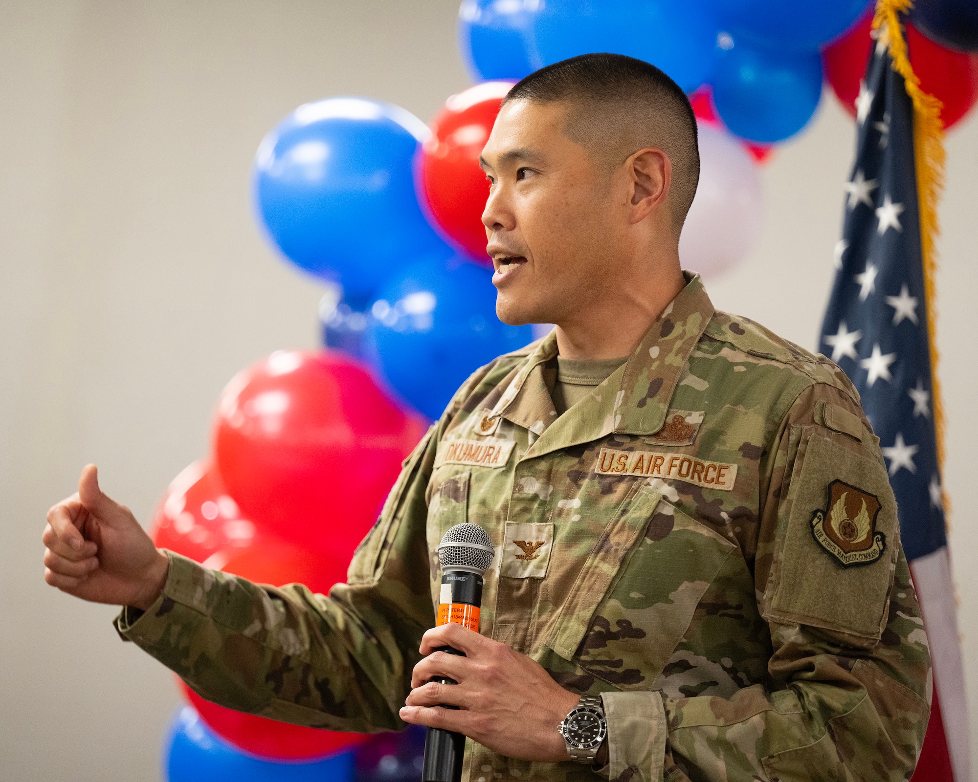 A man in camouflage uniform speaks holding a microphone. There are balloons behind him