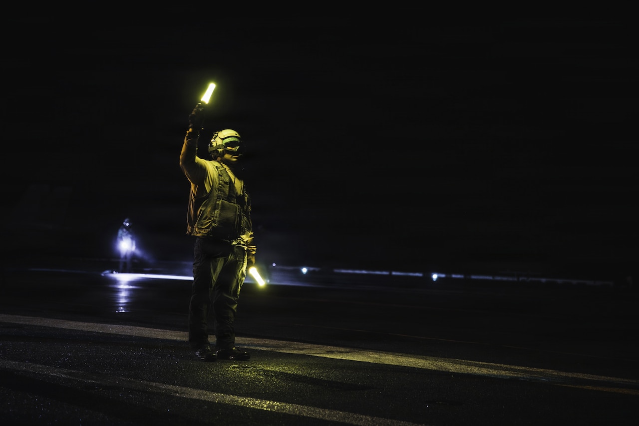 A person in a military uniform signals to an aircraft at night while aboard a military ship.