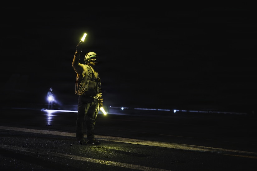 A person in a military uniform signals to an aircraft at night while aboard a military ship.