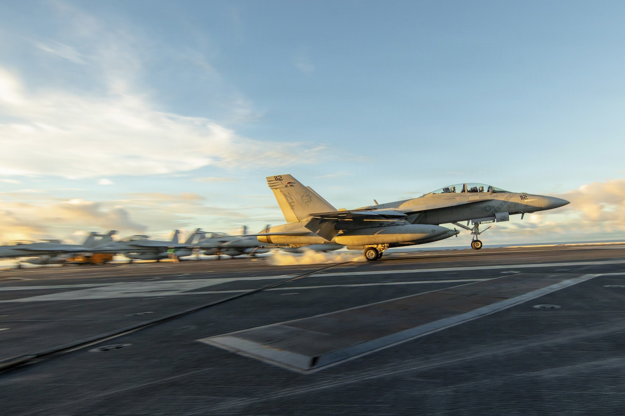 A military jet lands on the deck of an aircraft carrier during the day.