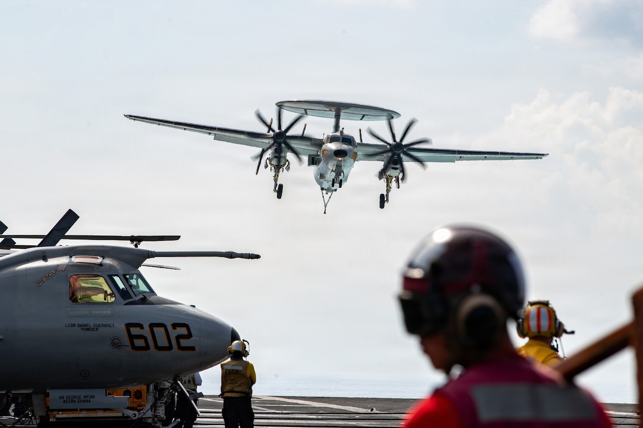 A military aircraft prepares to land on the flight deck of an aircraft carrier during the day as several people in military uniforms wait for the landing.