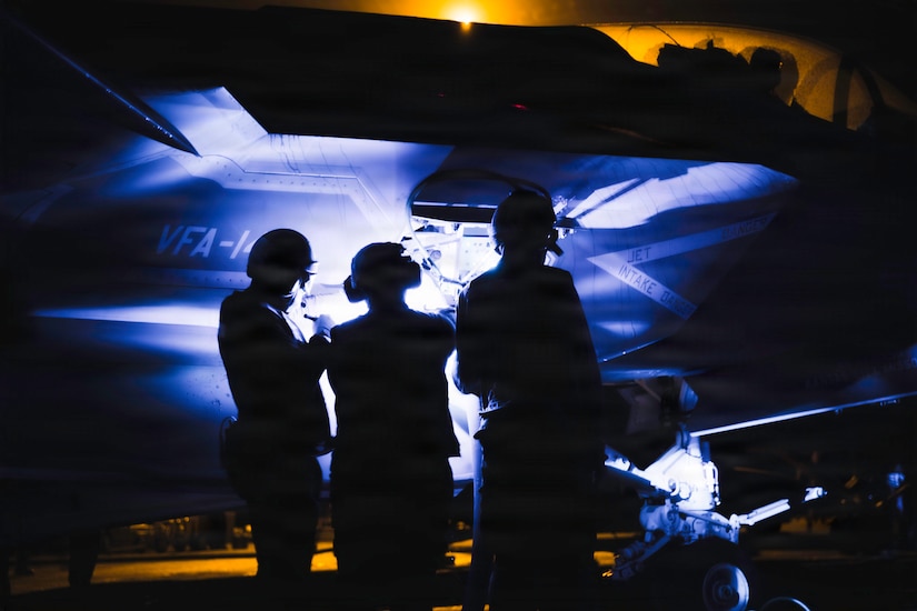 Three people in military uniforms check equipment on a military jet at night.