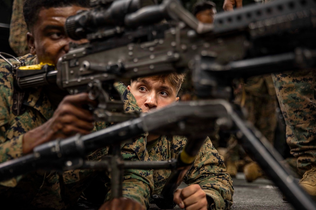A Marine looks through the scope of a weapon as a fellow Marine watches from the right in a dimly lit area.