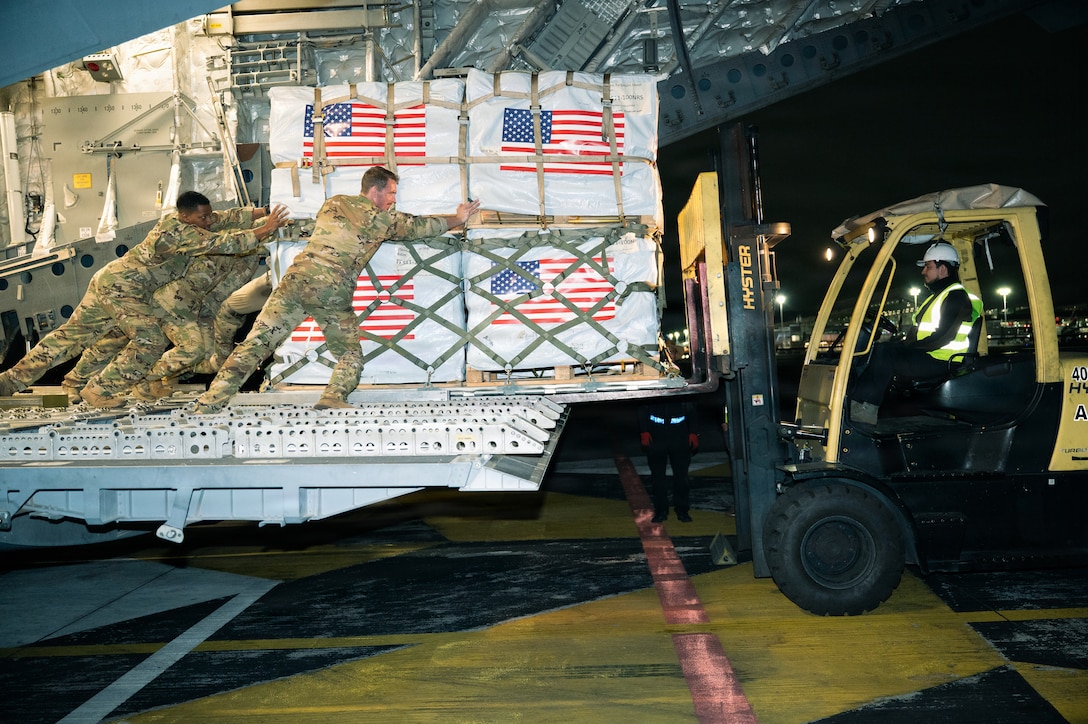 Airmen wearing camouflage uniforms push a pallet of cargo from the tailgate of a large aircraft onto a forklift driven by a man wearing a white helmet at night.