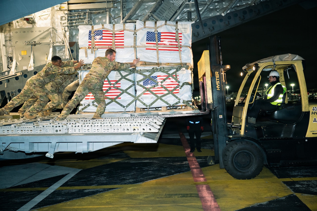 Airmen wearing camouflage uniforms push a pallet of cargo from the tailgate of a large aircraft onto a forklift driven by a man wearing a white helmet at night.