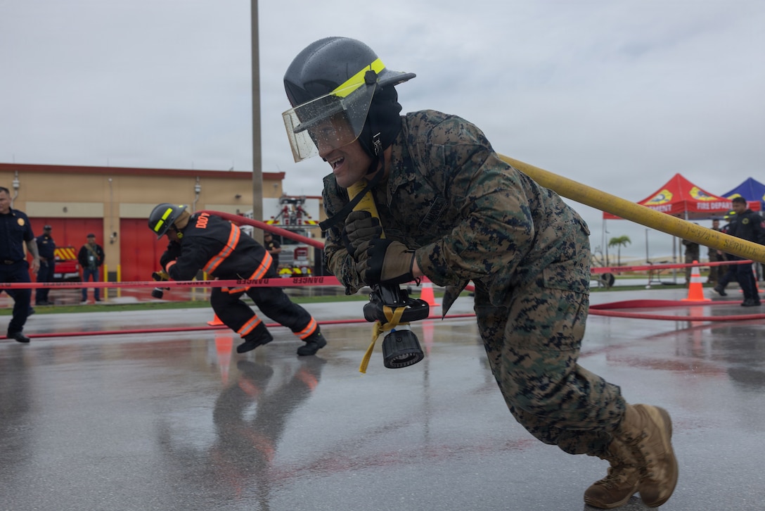 A Marine wearing a camouflage uniform and a firefighter's helmet pulls a firehose in a lane demarcated by red tape while a fireman in firefighting uniform is doing the same thing in another lane.
