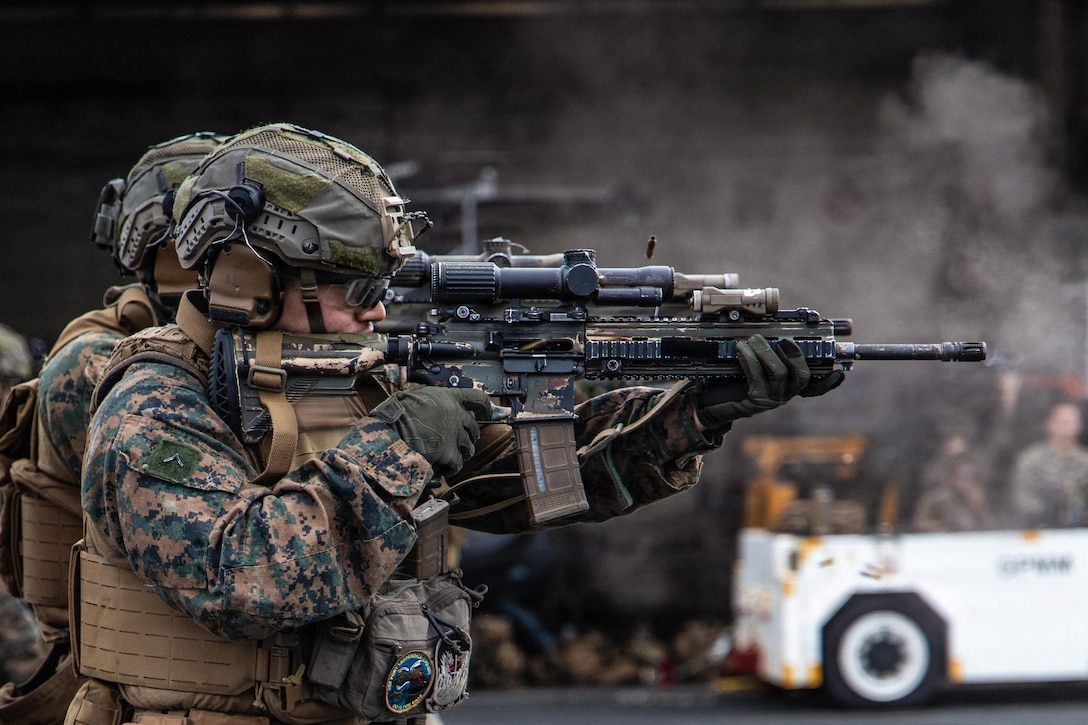 Two Marines wearing camouflage uniforms, tactical vests and helmets shoot rifles on the deck of a ship.