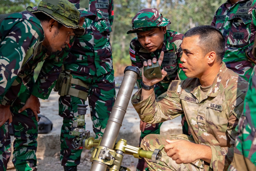 A U.S. soldier wearing a camouflage uniform kneels next to a mortar tube and talks to a group of Indonesian soldiers wearing camouflage uniforms.