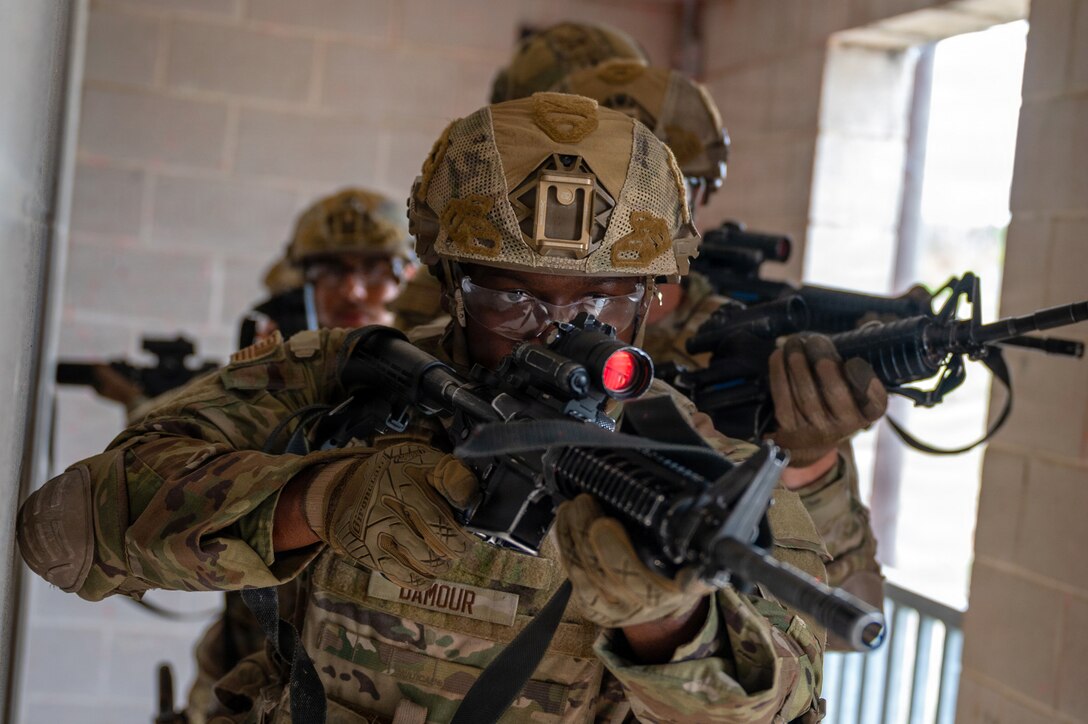 Airmen in helmets point weapons in different directions while walking in a line down a hallway.