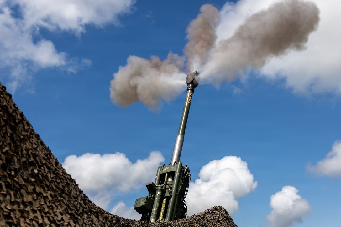 Three plumes of smoke shoot from a howitzer partly obscured by tent netting into blue, cloudy sky.