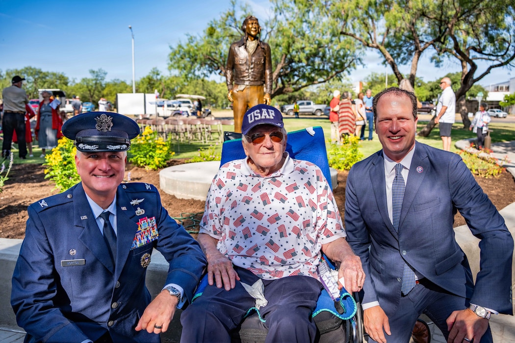 Three men sit and smile in front of a bronze statue