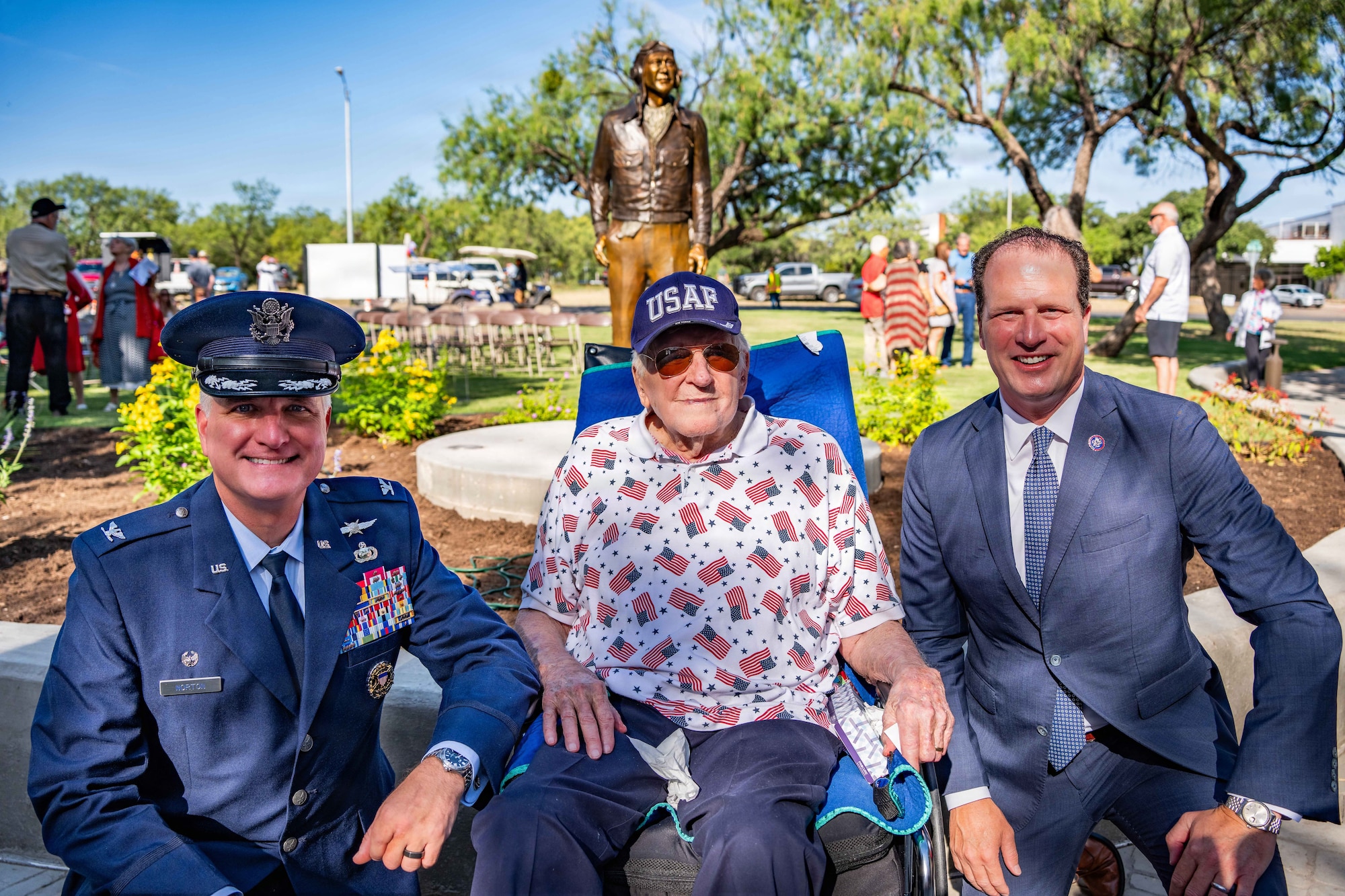 Three men sit and smile in front of a bronze statue