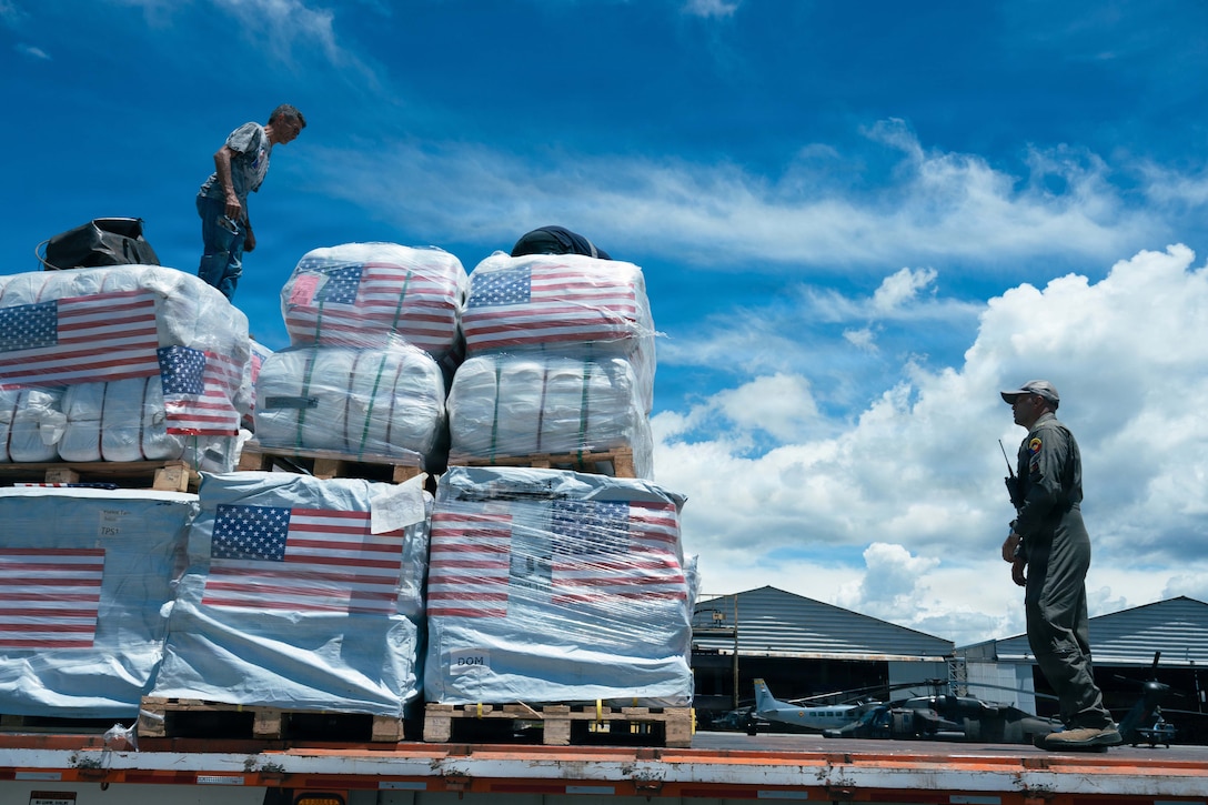 A service member stands on top of cargo that is sitting on a road, and another faces the cargo from the road. Aircraft, buildings and a blue sky are in the background.