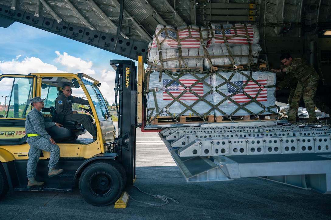 Two airmen on a yellow forklift offload cargo from an aircraft as another airman on the aircraft pushes the cargo. A blue sky and clouds are in the background.