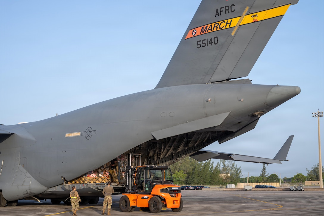 Two service members in camouflage uniforms stand next to a forklift and look into the back of a large military aircraft full of cargo.