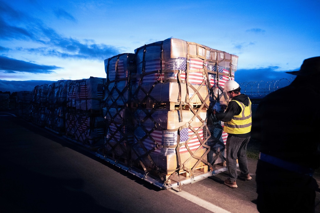 A service member wearing a yellow vest and white hard hat stands on a road in front of pallets of secured cargo at night. American flags are wrapped around the cargo packages.