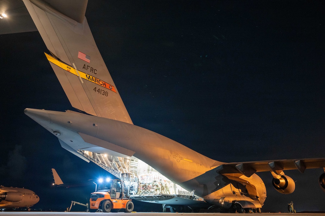 Service members load cargo into the back of a large military transport aircraft on a tarmac at night.