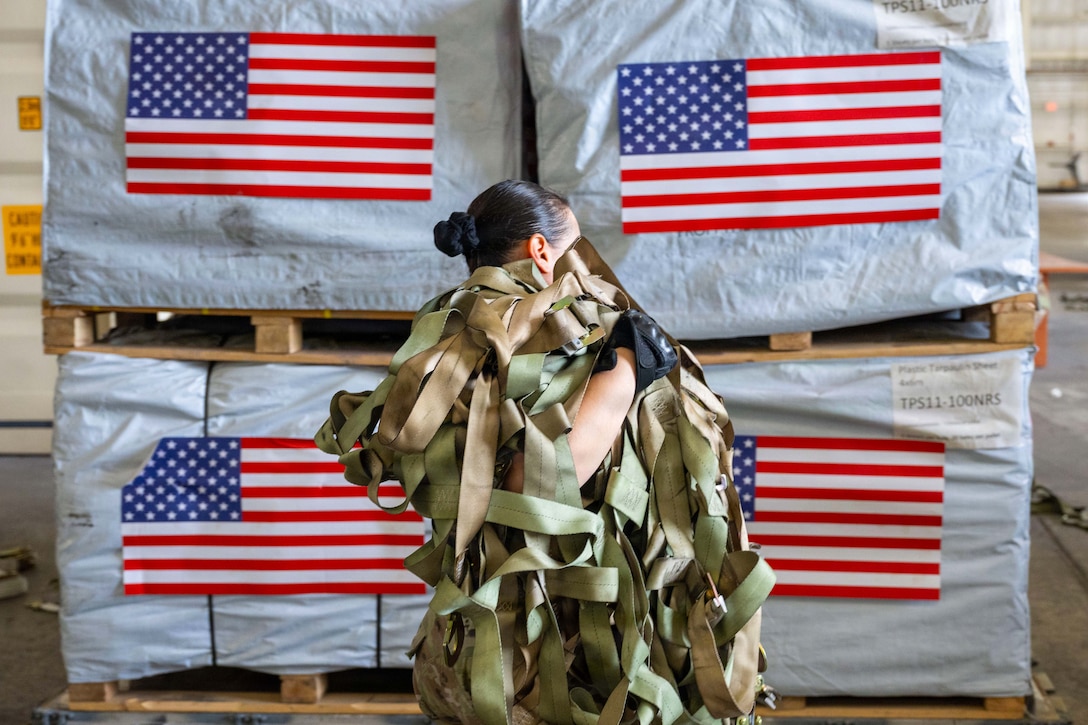 A service member in camouflage uniform holds cargo tiedowns in front of a pallet of cargo wrapped in white bags with an American flag on each bag.