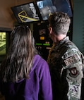 A U.S. Airmen stands with their spouse as they look at an engine in the viewing area of a test cell.