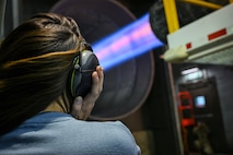 A person observes an engine run at full afterburner in a test cell. They are holding their earpotection over their ears and their hair is blowing from thw wind.