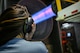 A person observes an engine run at full afterburner in a test cell. They are holding their earpotection over their ears and their hair is blowing from thw wind.