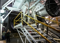 A maintenance Airman gestures for a tour group to move forward in a engine test cell.