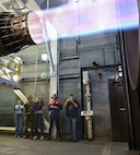 A group of people stand beneath a F15-E Strike Eagle Egine running at full afterburner in a test cell.