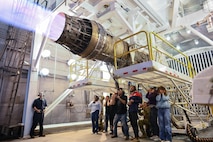 A group of people stand beneath a F15-E Strike Eagle Egine running at full afterburner in a test cell.