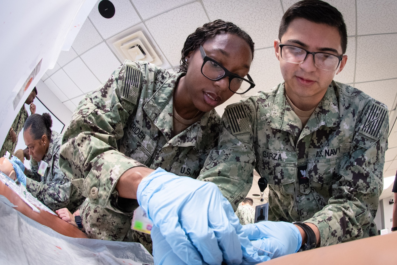 *Training*  Hospitalman Jamiyah Crider, center, coaches Hospital Corpsman 3rd Class Juan Garza, right, in training to insert an IV needle in a training aid during a skills fair conducted Thursday, August 20, 2026, at Navy Medicine Readiness and Training Command Cherry Point.  The Skills Fair evaluated the knowledge of corpsmen assigned to the facility and trained them on competencies and tasks expected of them in an operational environment.