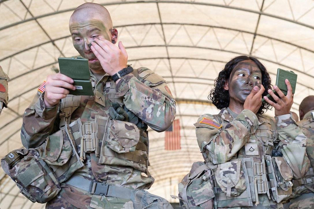 Two soldiers wearing camouflage uniforms apply camouflage face paint under a large canopy structure outdoors during daytime.