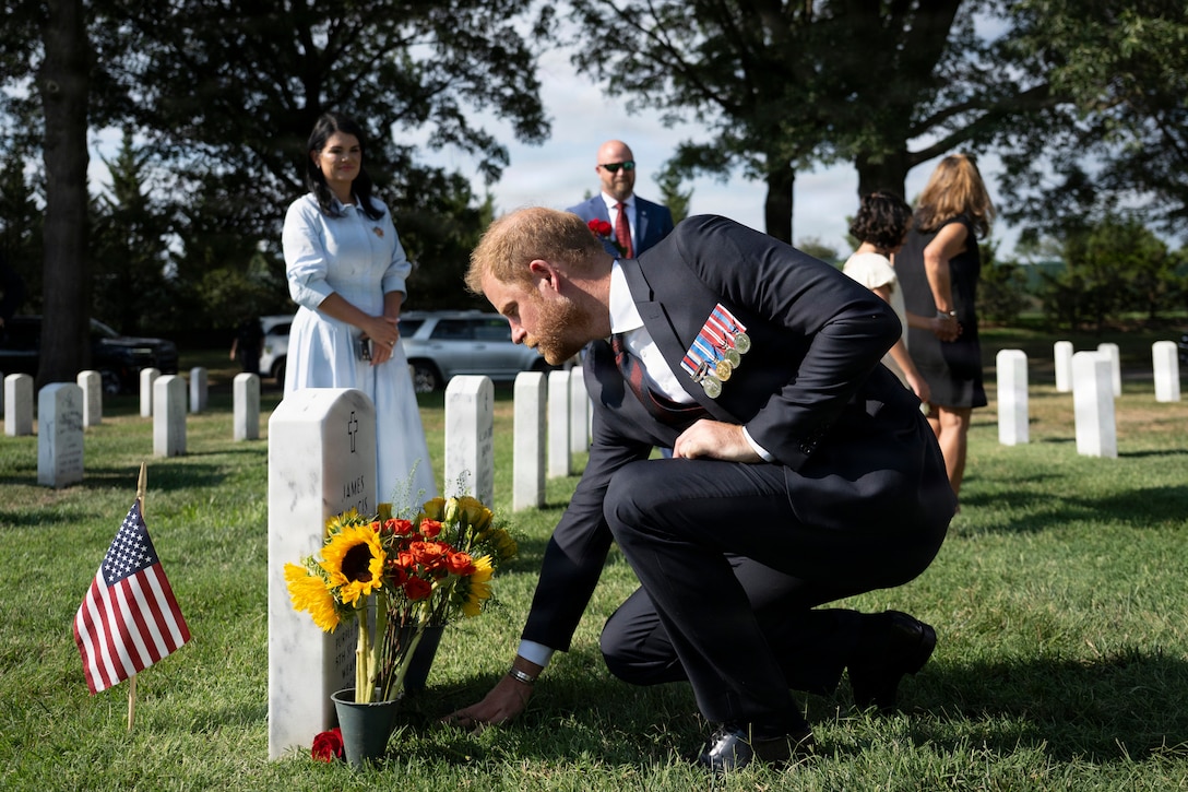 A man in a civilian suit with military medals affixed to his chest kneels before a gravestone in a cemetery during daytime.