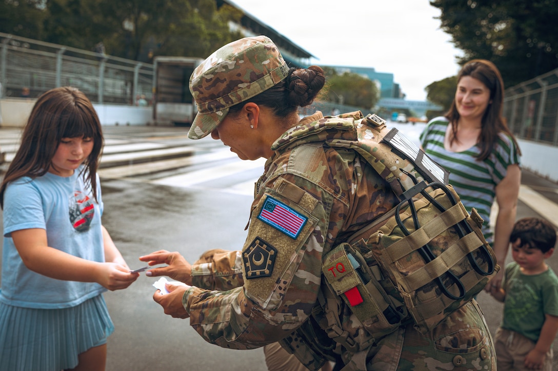 A soldier wearing a camouflage uniform and a tactical vest hands a sticker to a child while an adult and another child look on outside on a bridge during daytime.