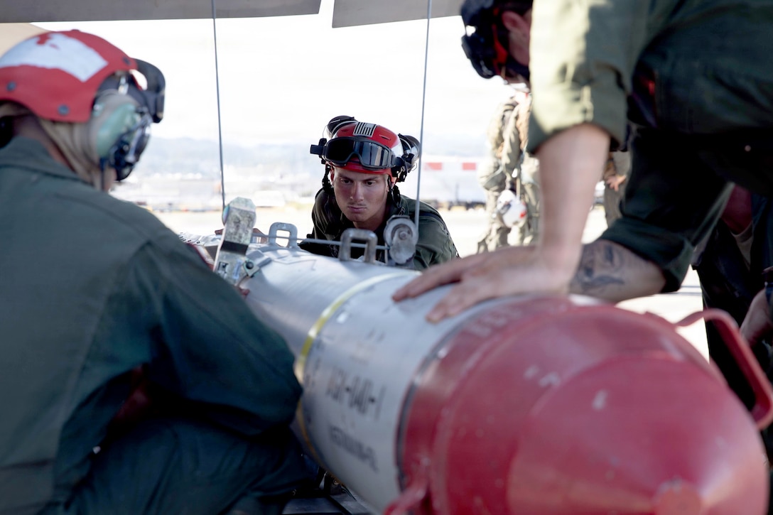 Three Marines wearing aviation jumpsuits load a missile onto a fighter jet in a hangar during daytime.