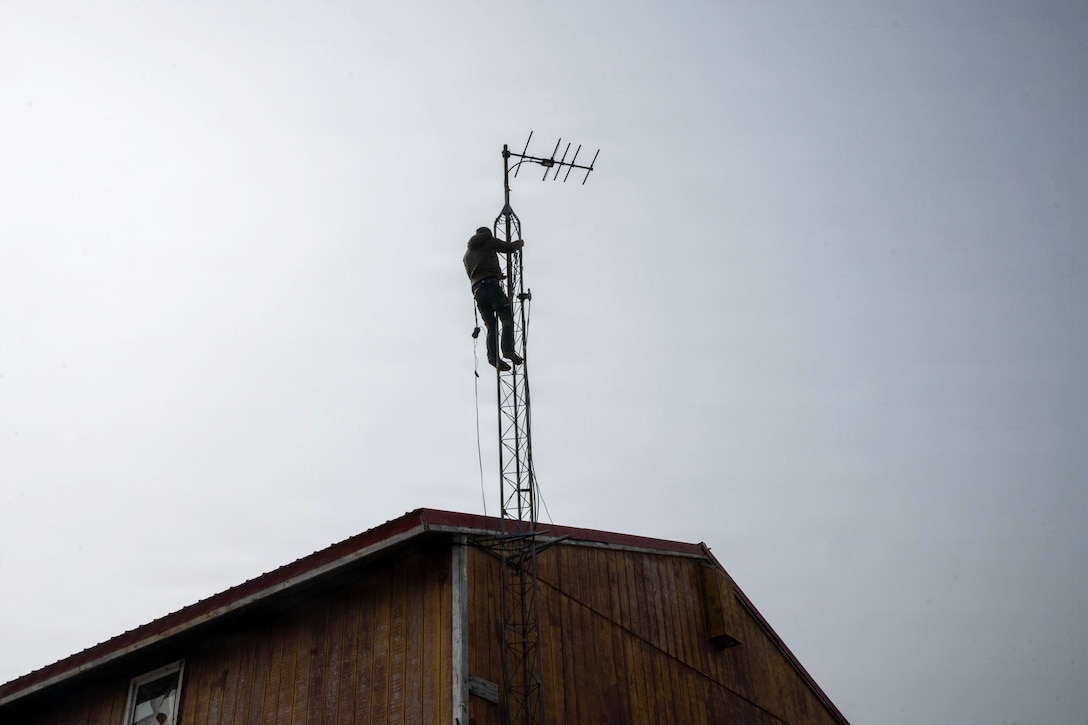 A service member climbs an antenna on the sloped roof of a building with wood siding on a gray day.
