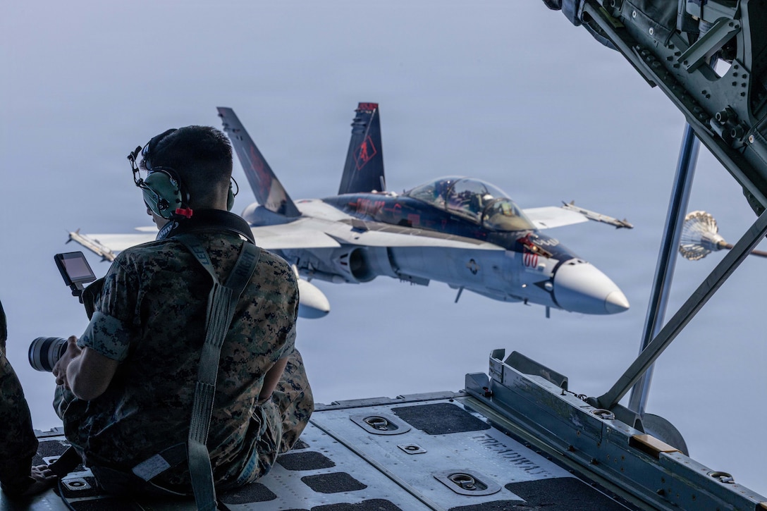 A Marine holds a video camera while sitting in the open hatch of an aircraft in the air diagonal to a white fighter jet and parallel to a refueler aircraft's nozzle.