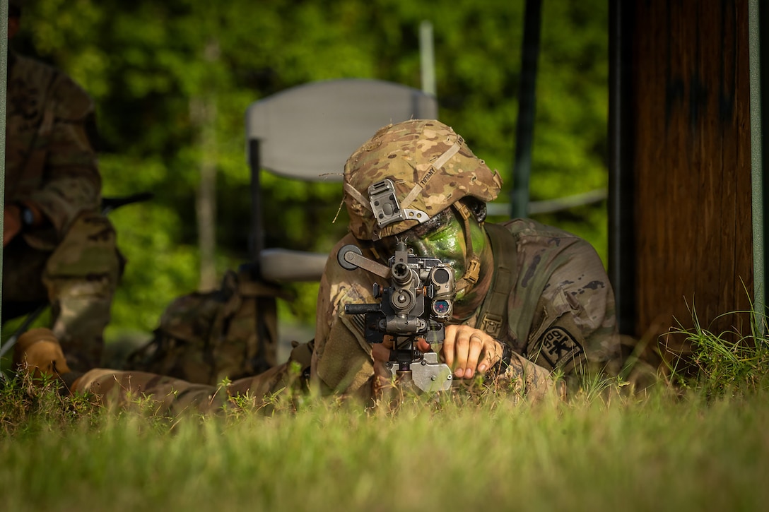 A soldier wearing green face paint lies on grass and looks through a piece of equipment that has four lenses.