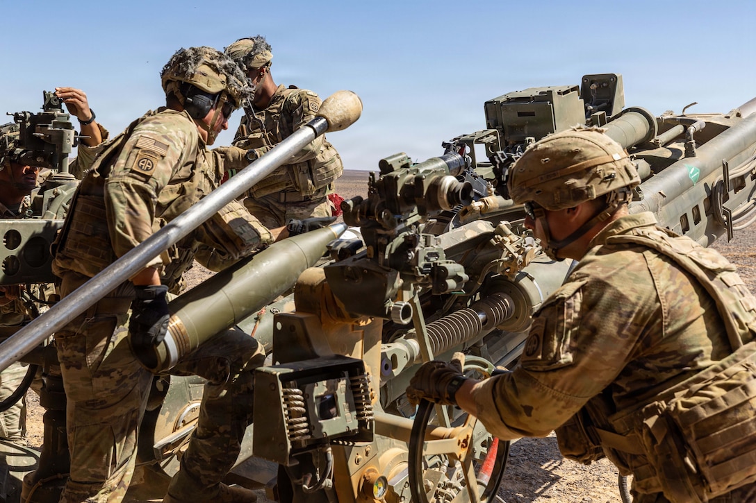 Four soldiers in camouflage uniforms operate a howitzer in the desert during the day.