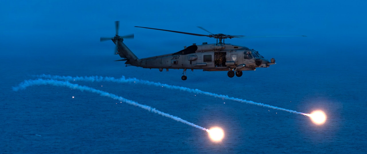 A helicopter flies at night over a body of water while deploying flares.