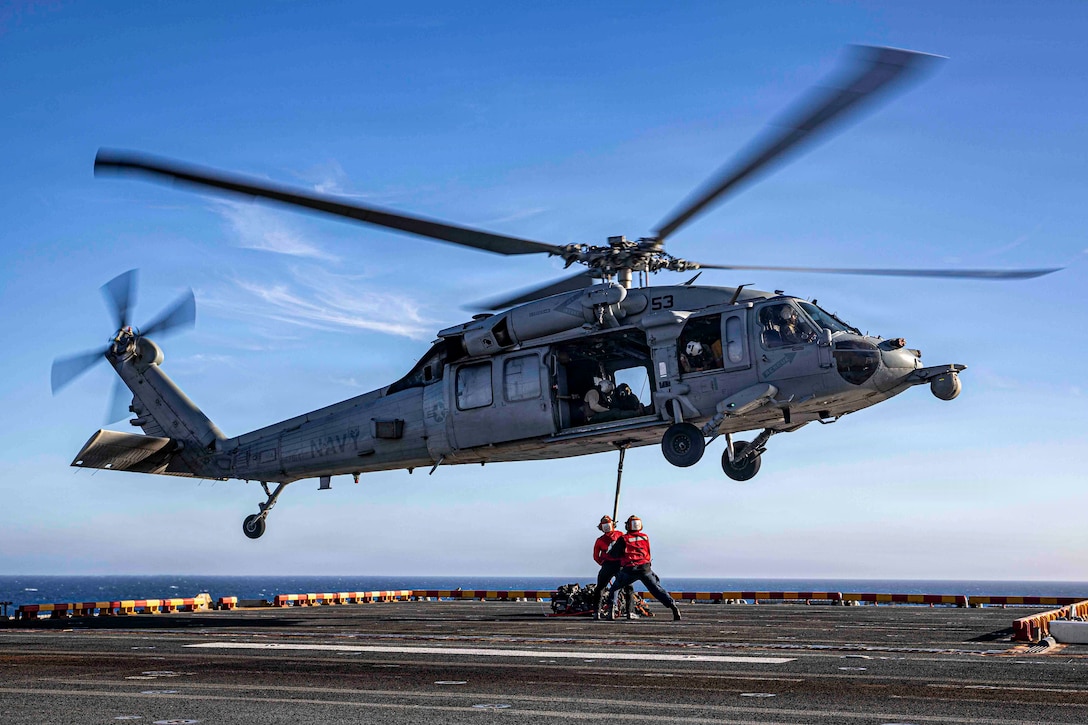 A helicopter hovers over the flight deck of an assault ship as two people wearing orange vests, green pants and head gear hold a device attached to the bottom of the aircraft.