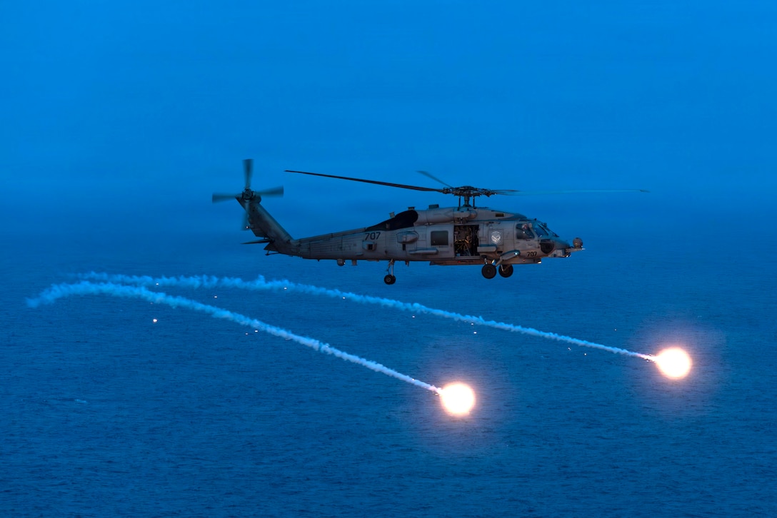 A helicopter flies at night over a body of water while deploying flares.