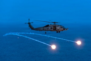 A helicopter flies at night over a body of water while deploying flares.