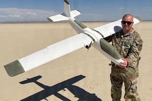 A man dressed in a camouflage military uniform holds a small white aircraft drone.