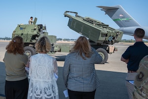 A group of people look at a combat vehicle with a plane in the background.