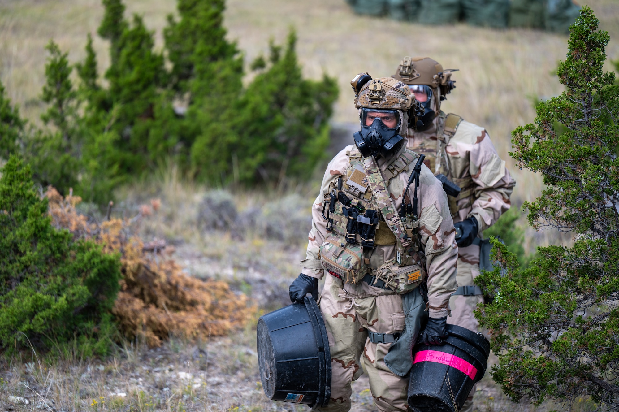U.S. Air Force Senior Airman Mitchell Massa, 366th Civil Engineer Squadron (CES) explosive ordnance disposal (EOD) technician, front, and Airman 1st Class Levi Womack, 341st CES EOD technician, approach a simulated chemical explosive device during an exercise at Limestone Hills Training Area, Montana, Aug. 18, 2026. Working in chemical protective gear adds heat, restricted mobility and limited visibility, conditions technicians must train in to stay effective against contaminated threats. (U.S. Air Force photo by Tech. Sgt. Aaron Thomasson)