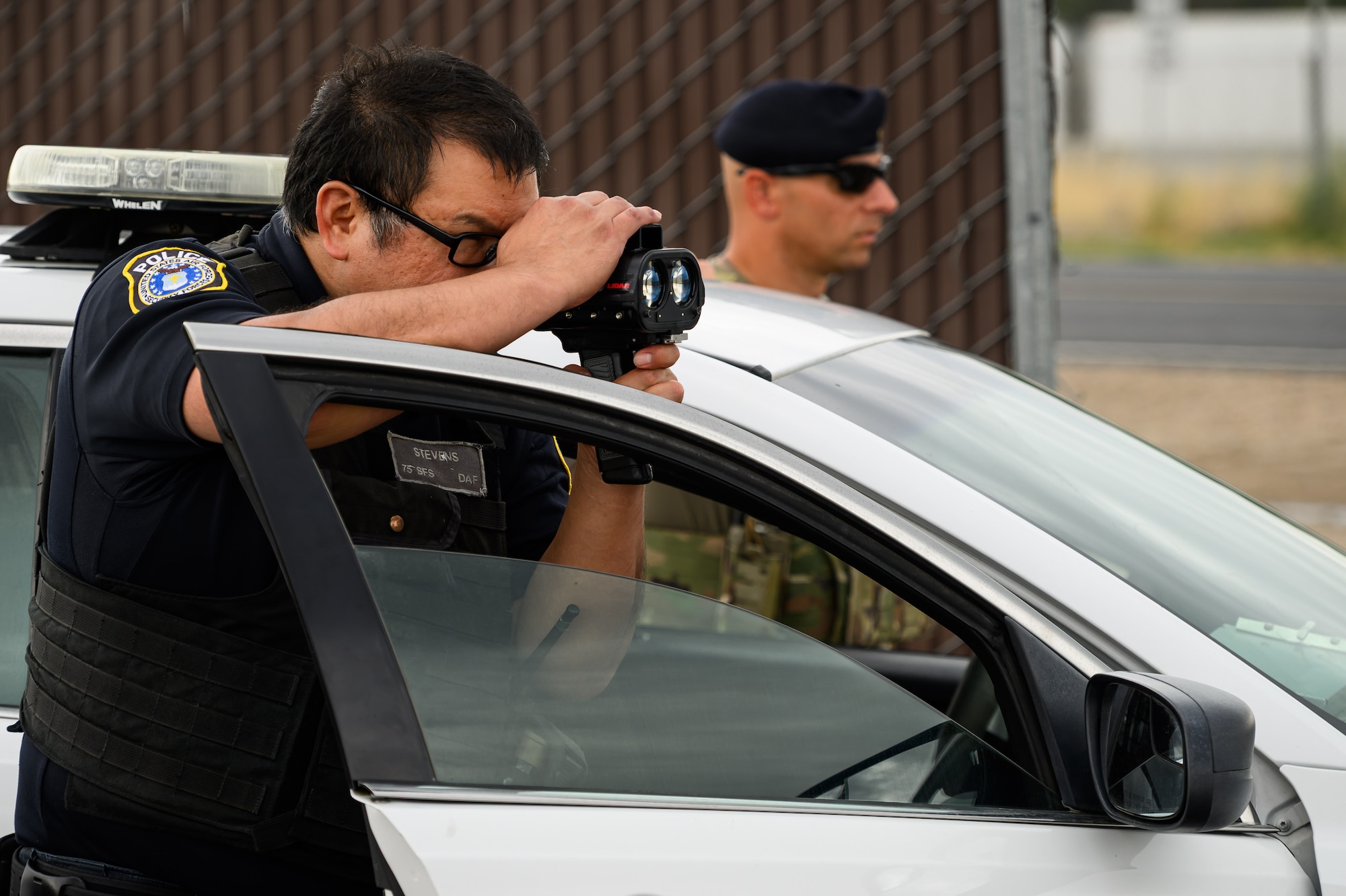 Two 75th Security Forces Squadron defenders stand just outside the doors of their patrol car during a speed enforcement operation. The defender in the foreground is looking through a radar device.