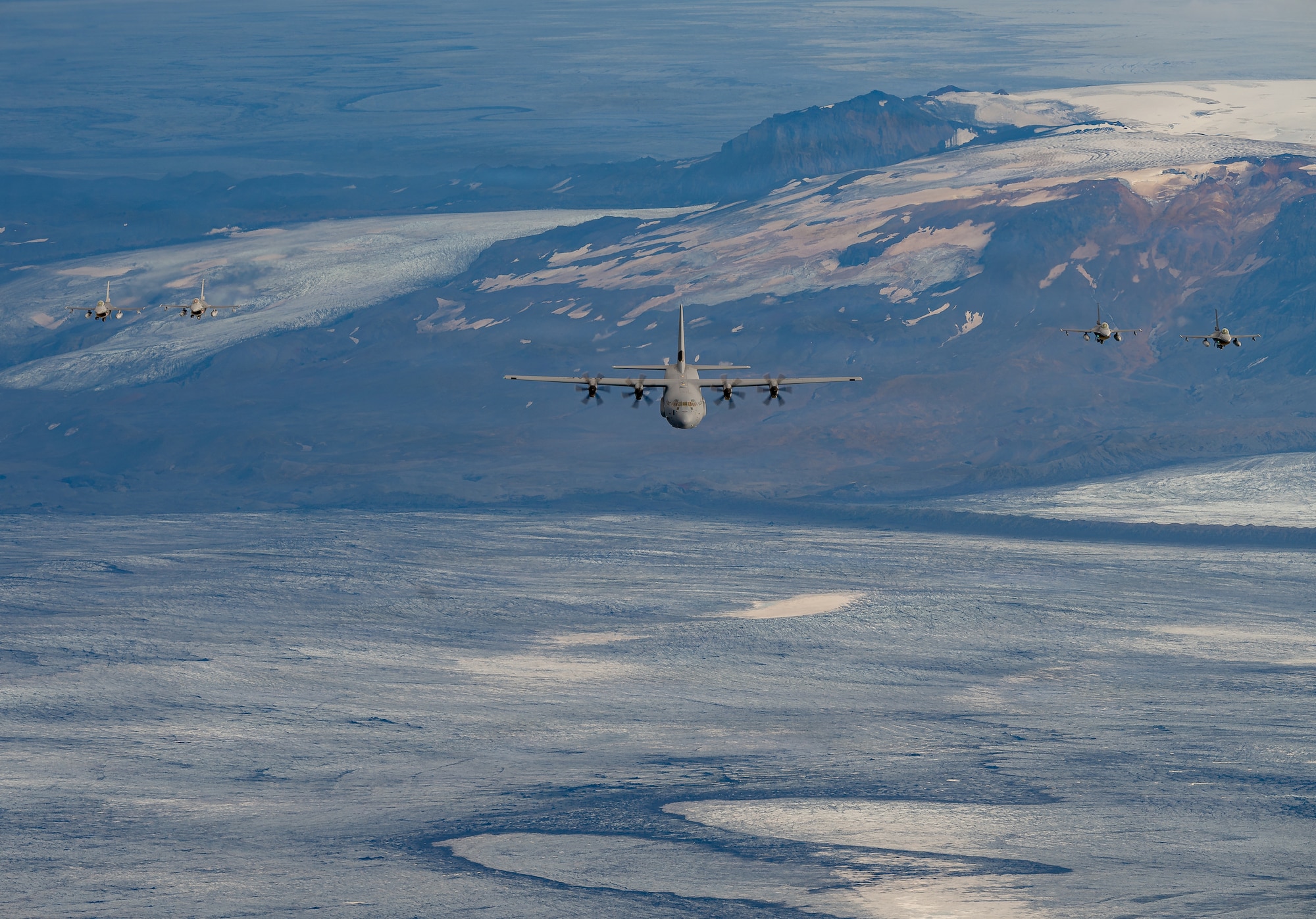 Four Belgian Air Force F-16 Fighting Falcon aircraft fly in formation with a U.S. Air Force C-130J Super Hercules aircraft assigned to the 37th Airlift Squadron above glacier-covered terrain in Iceland during exercise Northern Viking 26